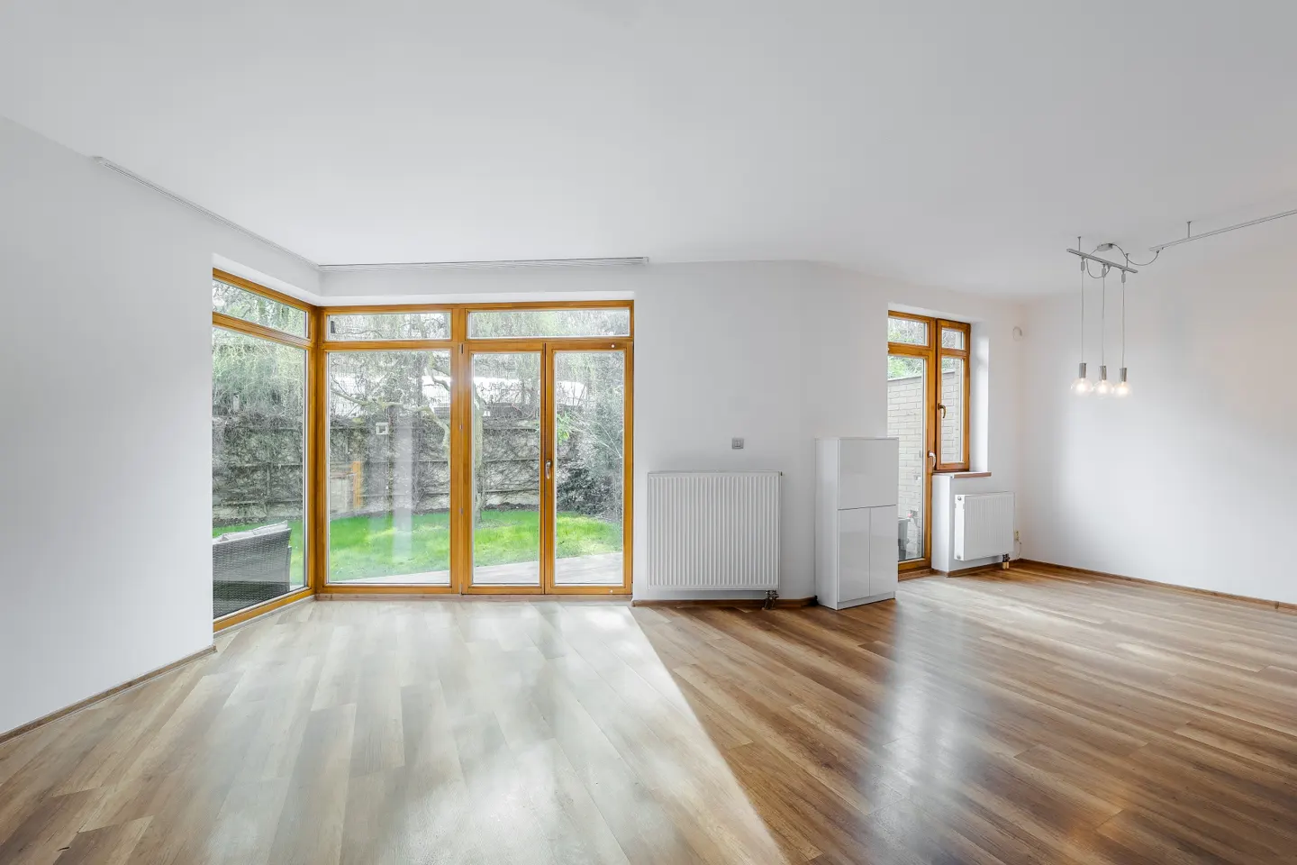 Bright, empty room with hardwood floors, white walls, and large windows overlooking a green yard. Radiators and a modern light fixture are visible.