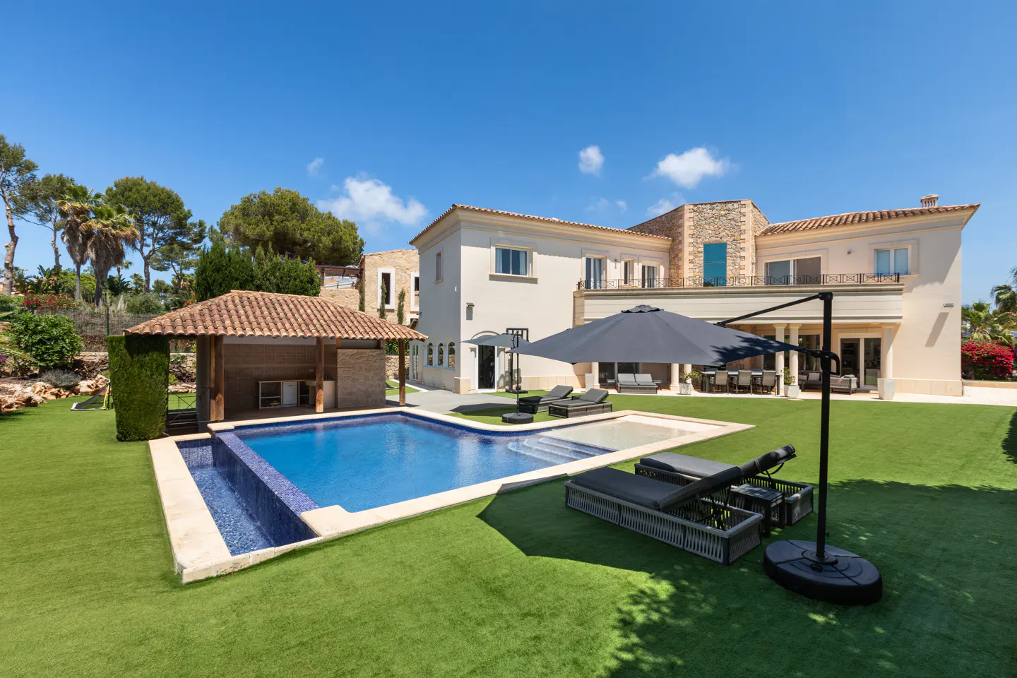 Luxury home exterior with a blue pool, cabana, lounge chairs, and a large umbrella on a green lawn under a blue sky.