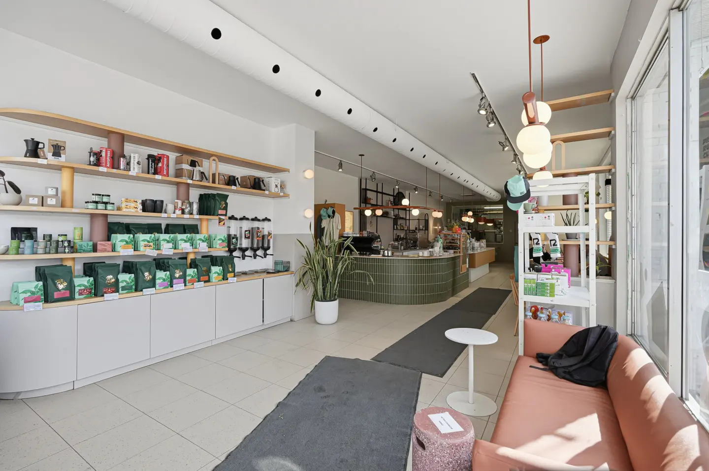 Bright, modern coffee shop interior with shelves of coffee beans, a green tiled counter, and a pink sofa near the window.