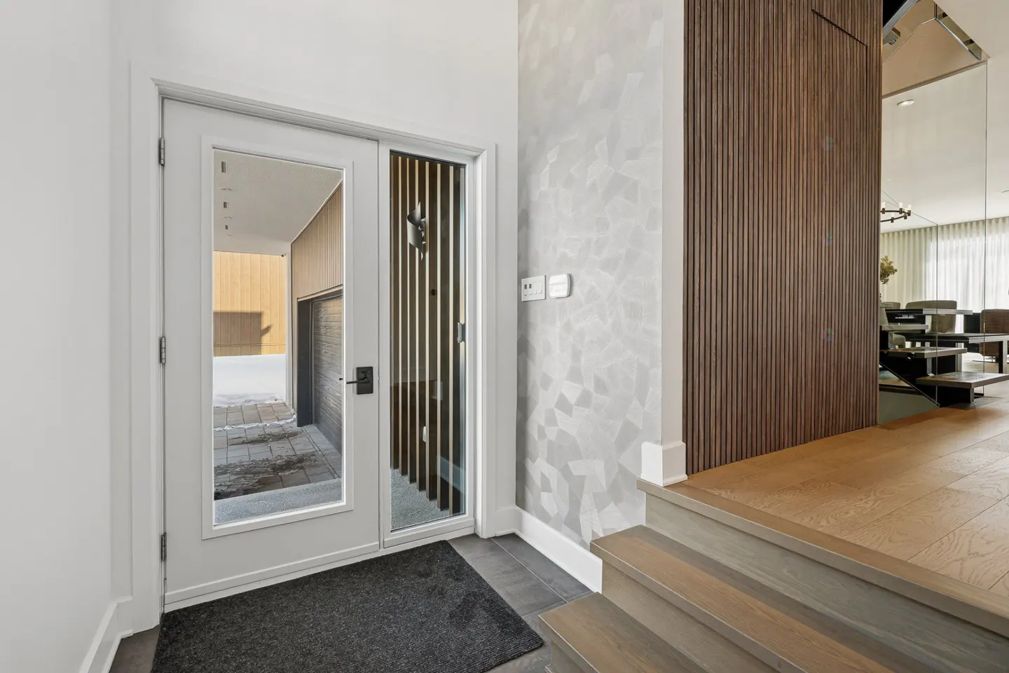 Modern home entryway with a white door, dark gray mat, and light gray walls with a geometric pattern. Wood stairs lead to a wood floor.