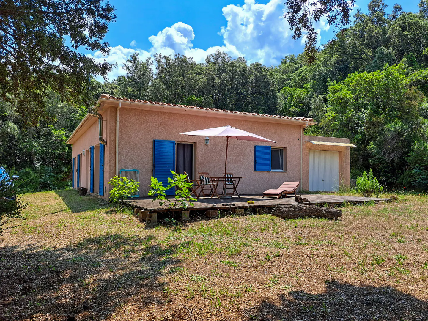 Exterior view of a one-story peach house with blue shutters, a patio with furniture, and a garage, surrounded by trees.