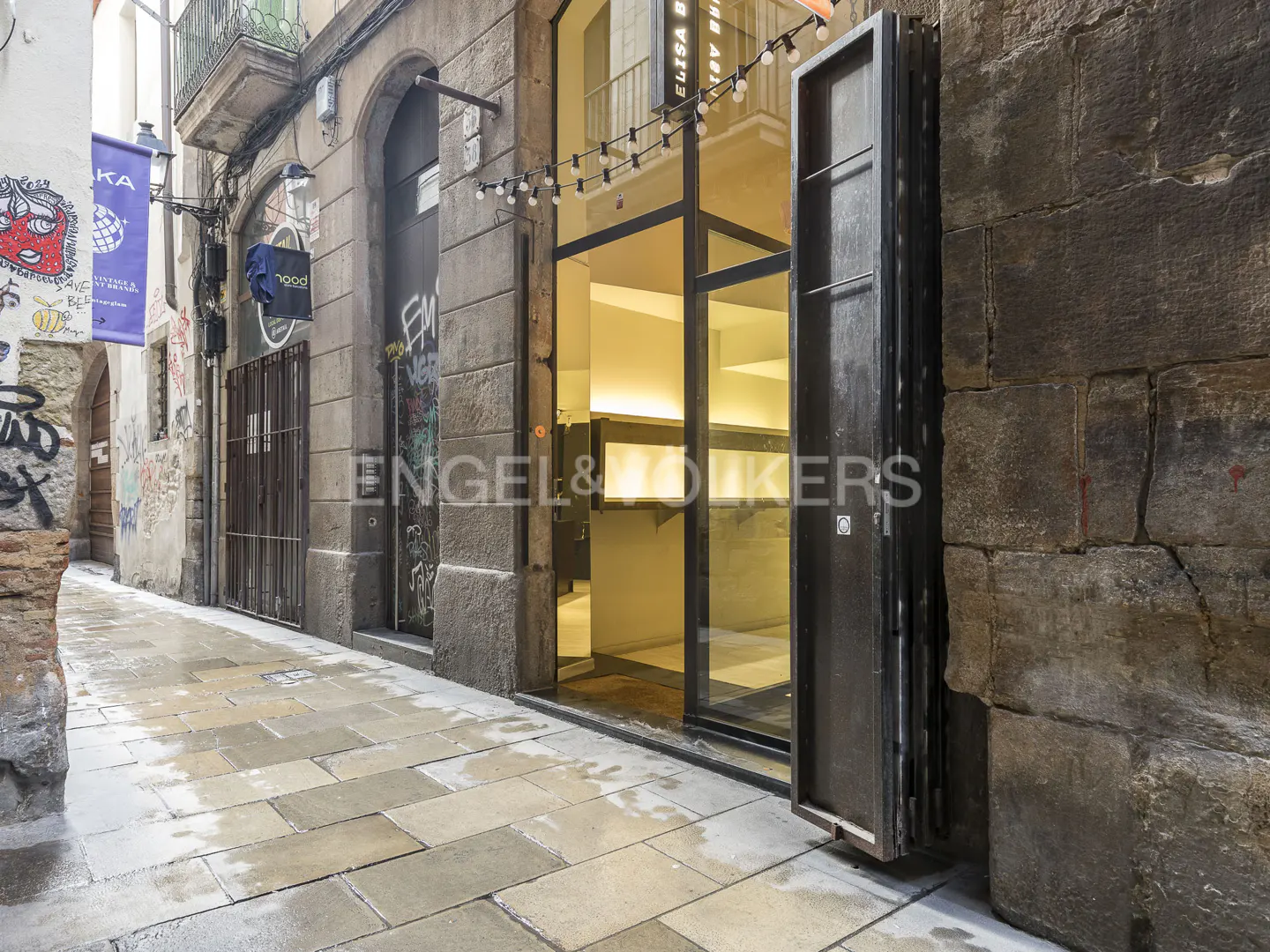 Engel & Volkers office on a narrow street with stone buildings and graffiti. The entrance has a black frame and string lights.