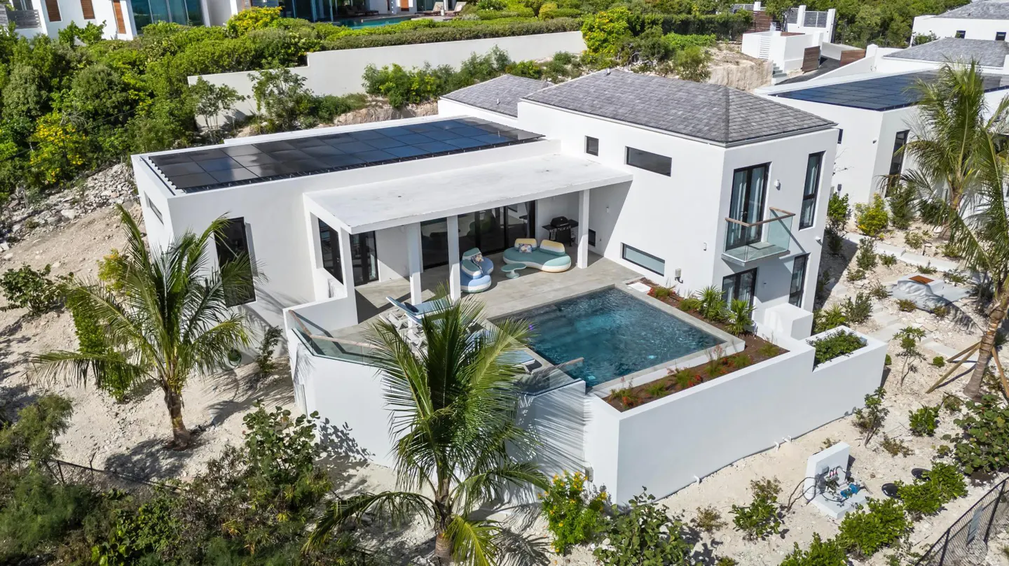 Aerial view of a modern white house with a pool, solar panels, and lush green landscaping.