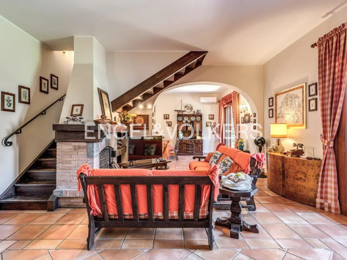 Living room with terracotta tile floor, orange sofas, fireplace, and staircase. Archway leads to another room. Engel & Volkers logo visible.