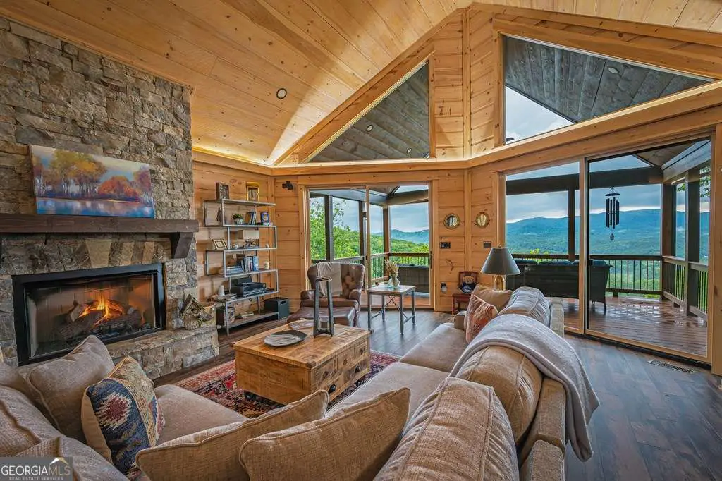 Cozy living room with stone fireplace, wood walls, vaulted ceiling, and large windows overlooking mountain views.