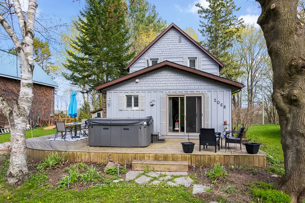 Exterior view of a light blue house with a hot tub on a wooden deck, surrounded by trees and greenery.