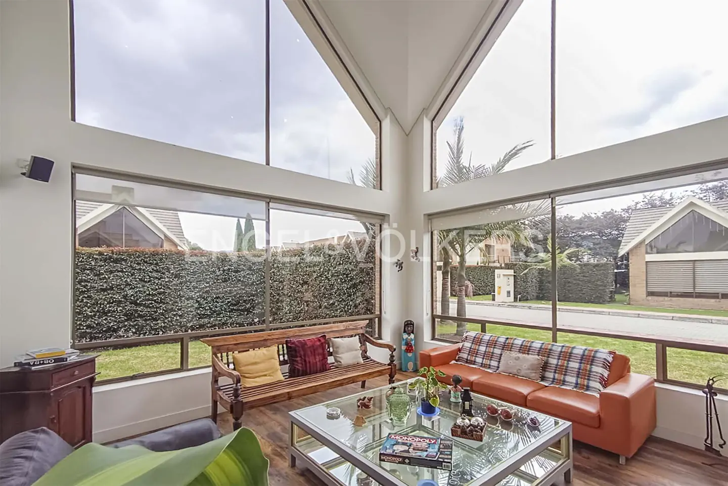 Bright living room with large windows showing green hedges and houses. A wooden bench and orange sofa face a glass coffee table.