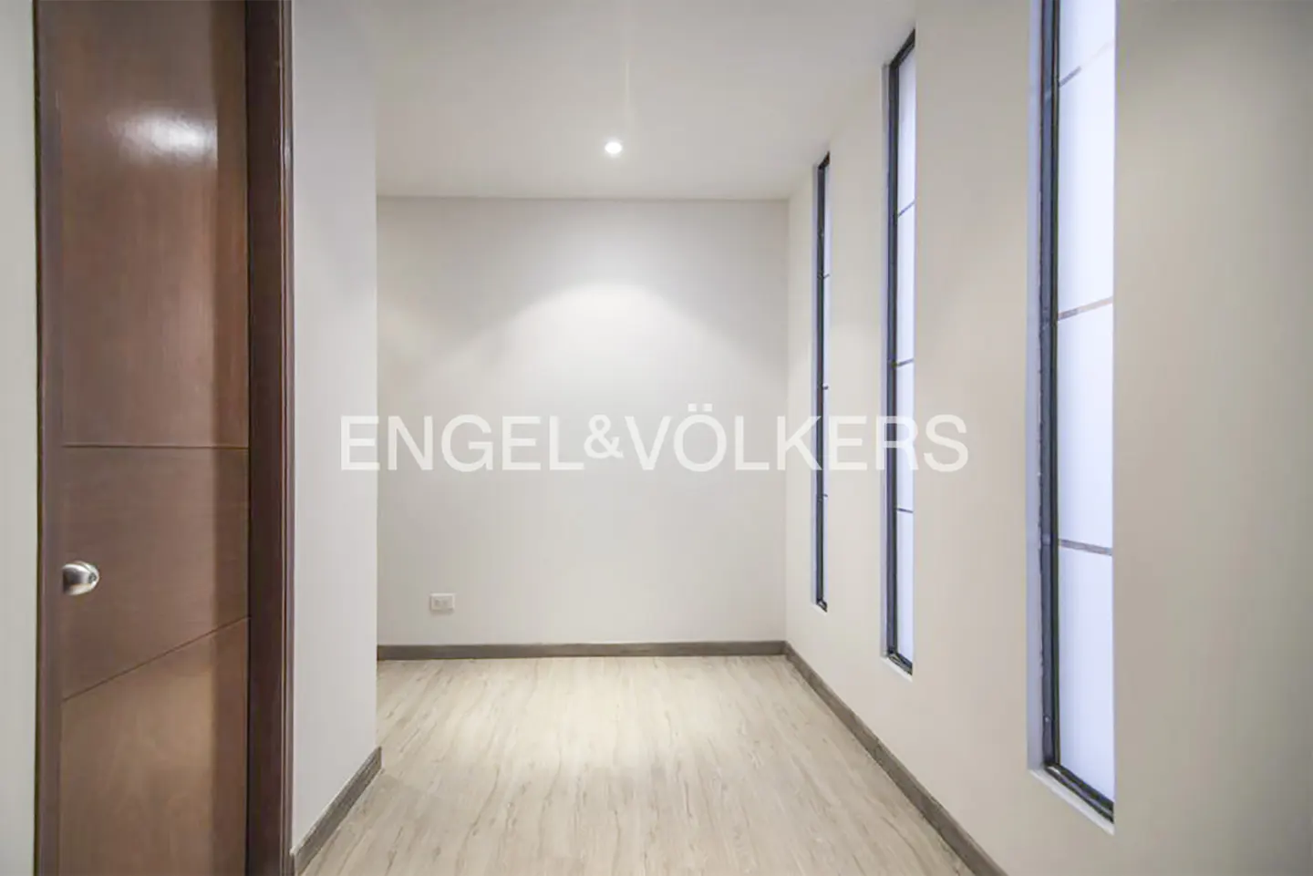 A bright hallway with light wood floors, white walls, a brown wooden door, and three tall, narrow vertical windows.