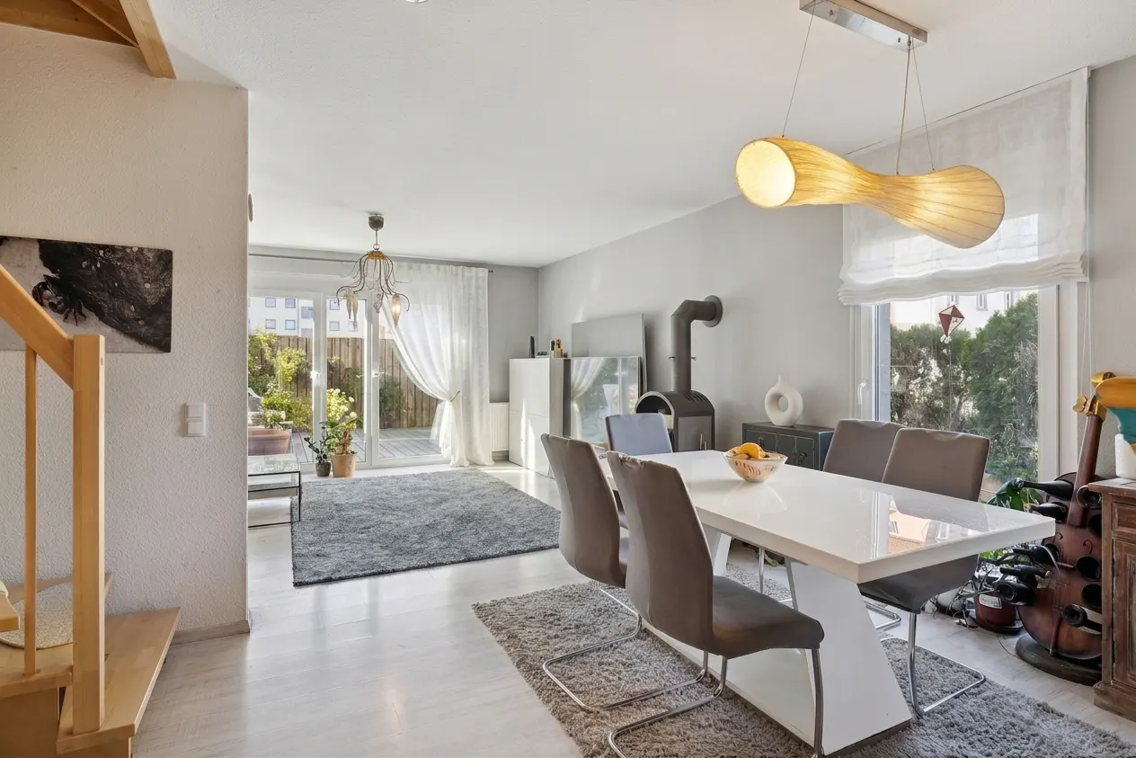 Bright, modern dining room with a white table, gray chairs, and a unique light fixture. A wood-burning stove and outdoor patio are visible.