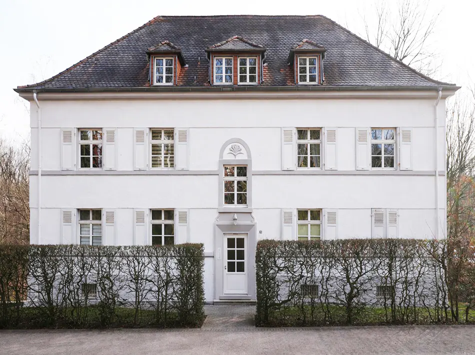 Two-story white house with a dark roof and white shutters, framed by a trimmed hedge.