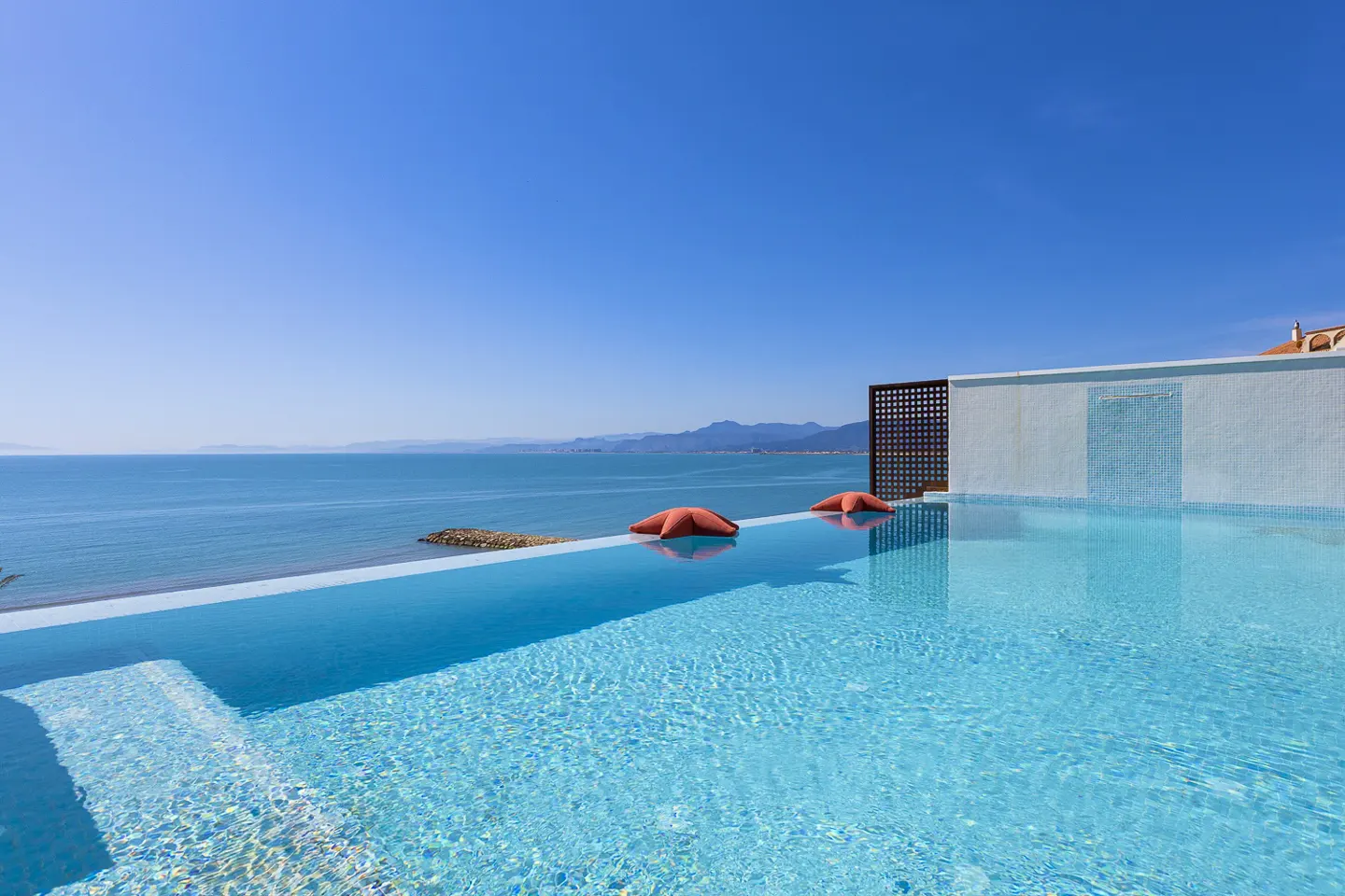 Infinity pool with two orange starfish floats overlooking the ocean under a clear blue sky.