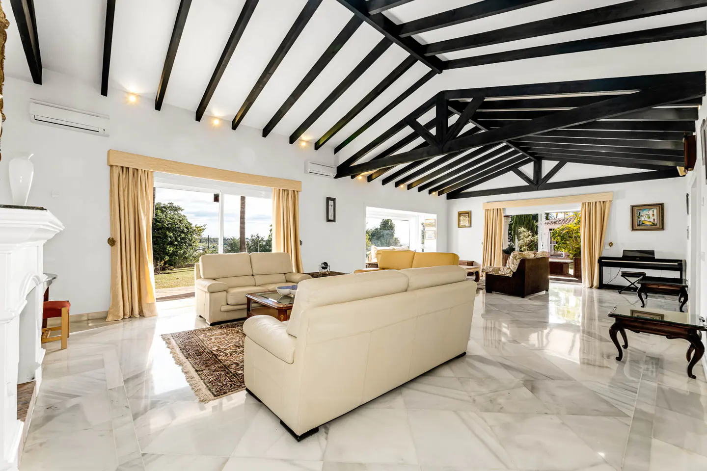 Bright living room with white walls, marble floors, and black beamed ceiling. Beige sofas face large windows with tan curtains.
