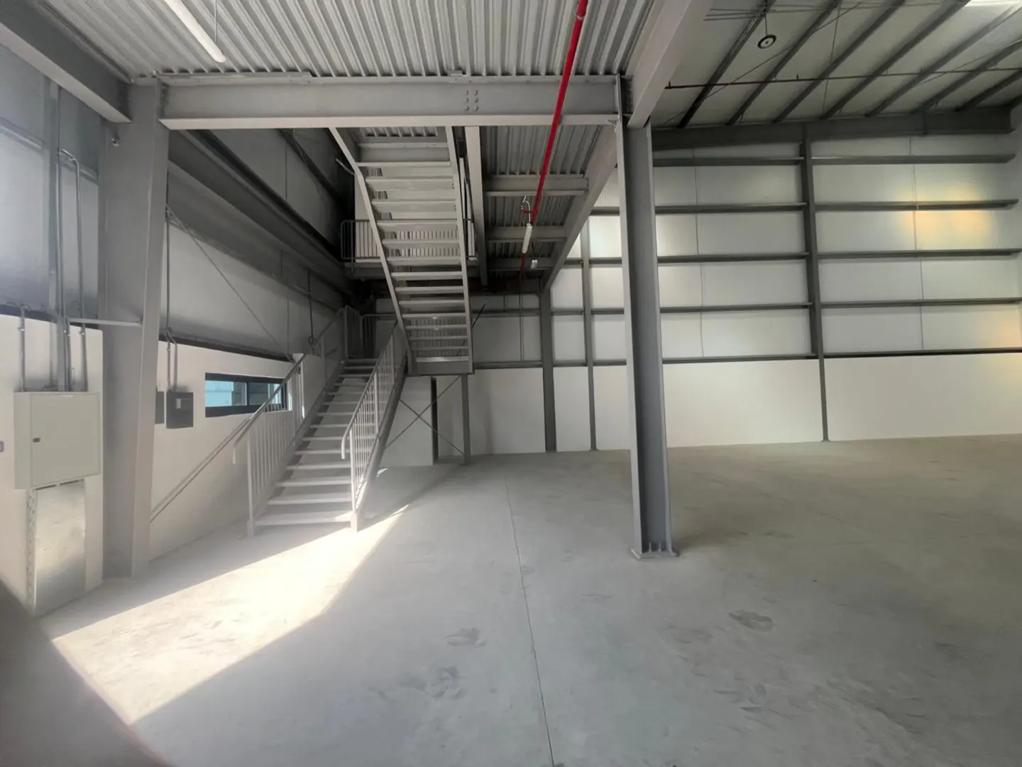 Interior of a large, empty warehouse with metal stairs leading to a second level. The walls and floors are gray.