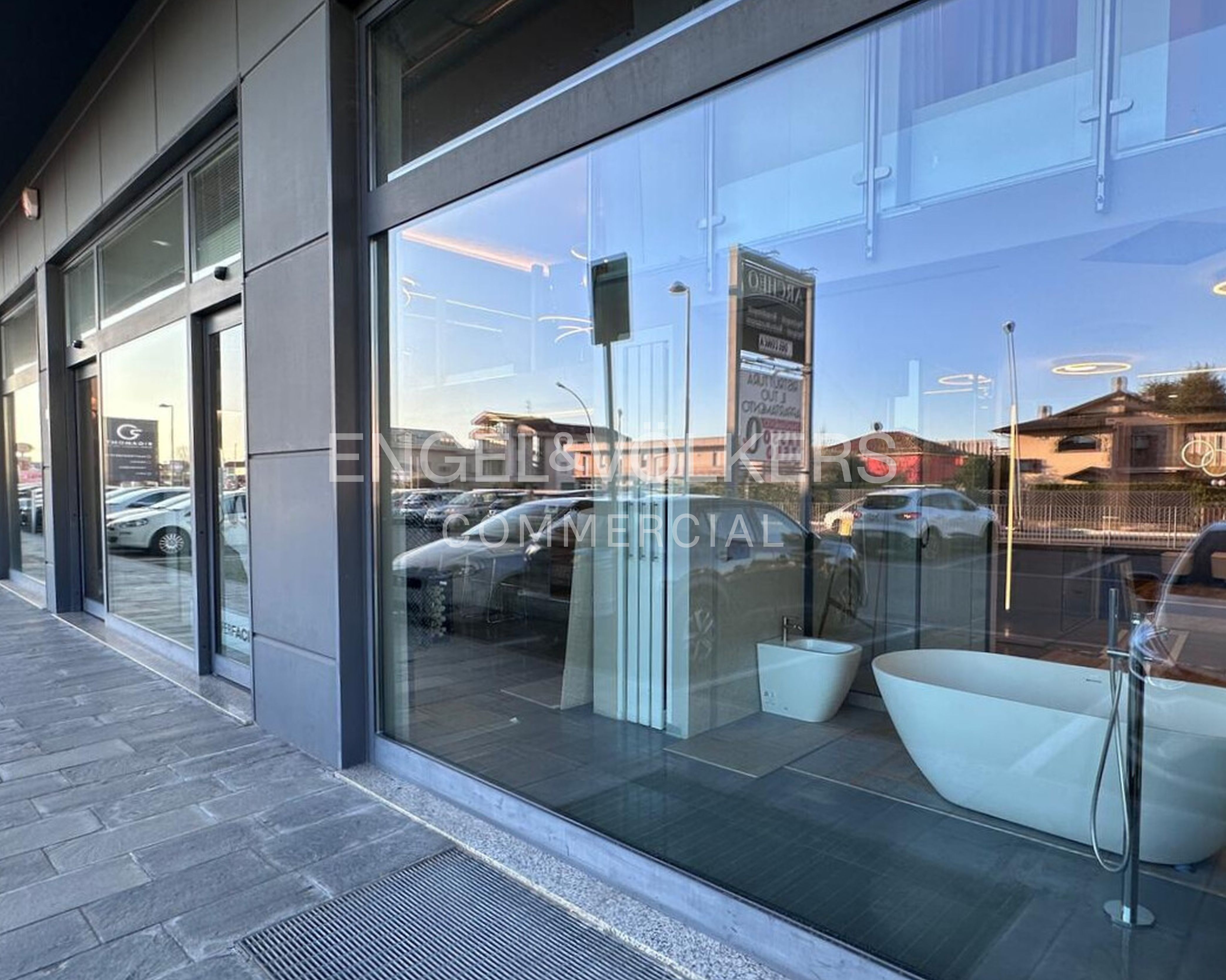 Modern showroom exterior with large glass windows displaying a white freestanding bathtub and bidet, reflecting the street.