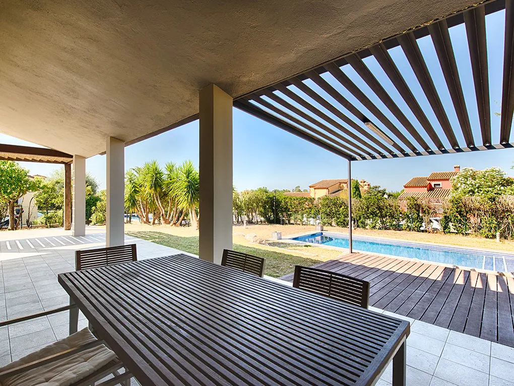 Outdoor patio with a wooden table and chairs, overlooking a pool and green lawn. A pergola provides shade.