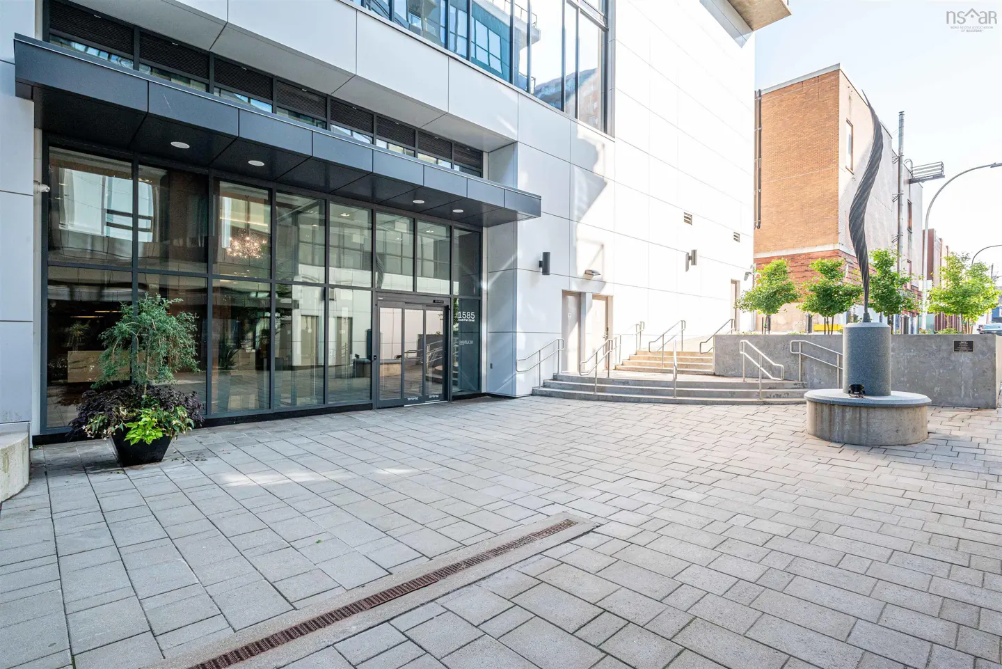 Modern building exterior with glass doors, a gray stone walkway, and a decorative fountain with trees.