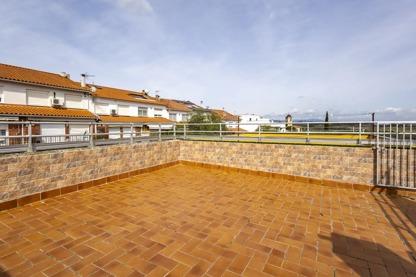Terracotta-tiled patio with brick wall and metal railing. White houses with orange roofs in the background under a blue sky.