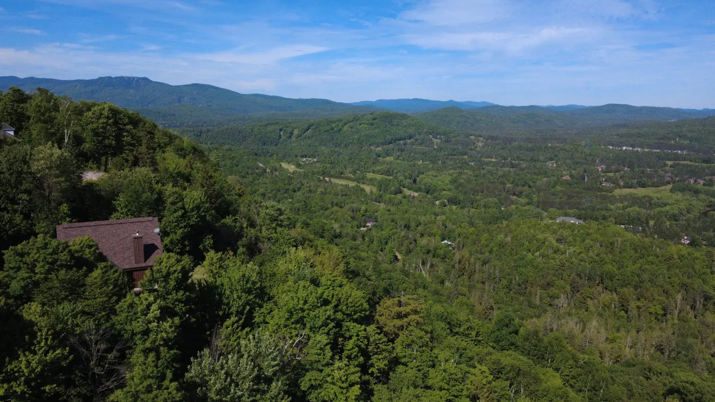 Aerial view of a brown house nestled in a lush green forest overlooking a vast mountain valley under a blue sky.