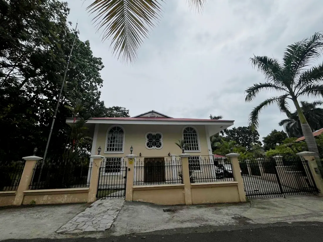Beige two-story house with a red roof, black iron fence, and palm trees against a cloudy sky.
