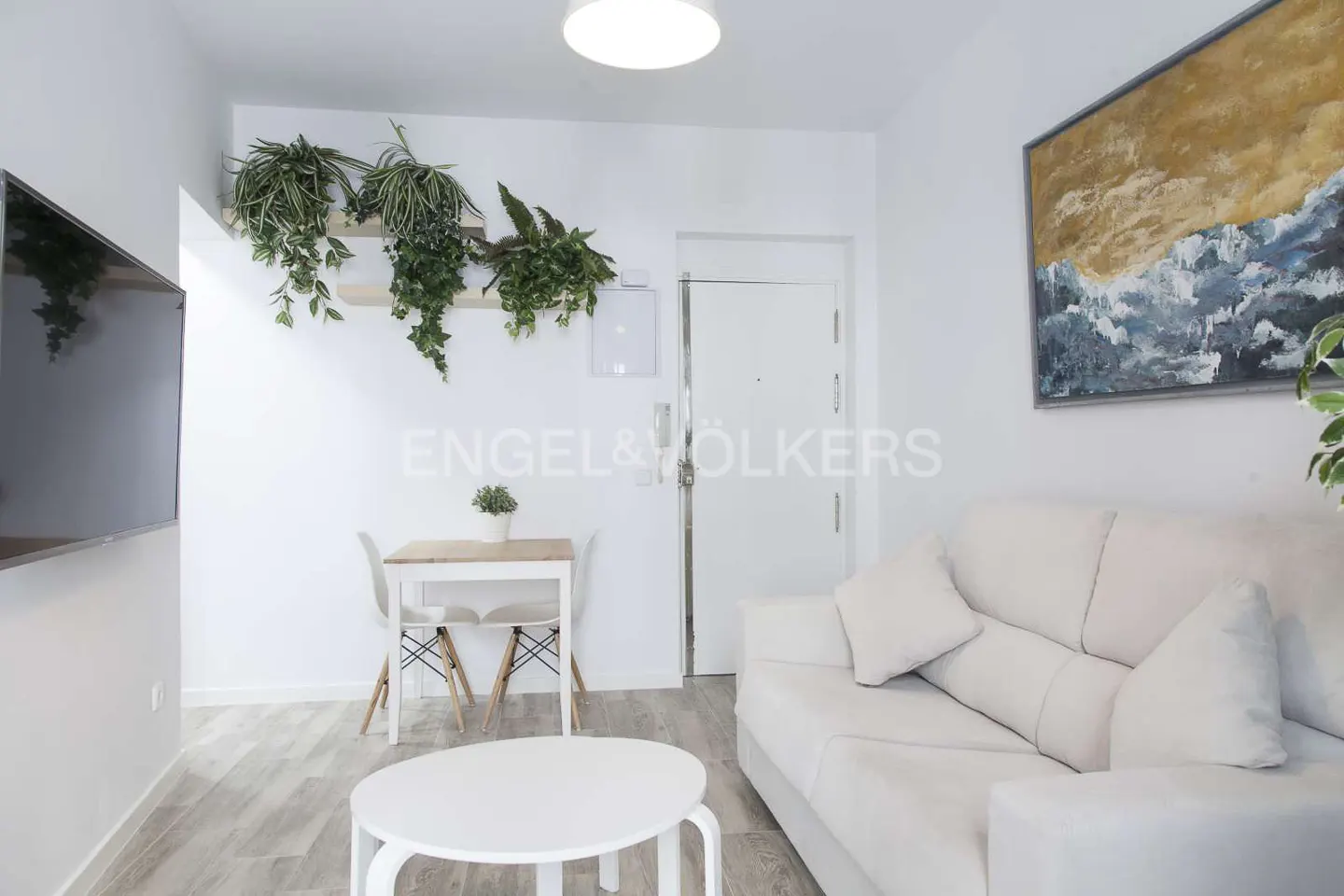 Bright living room with white walls, light wood floors, and white furniture. A painting hangs above a white sofa. Plants decorate the wall.