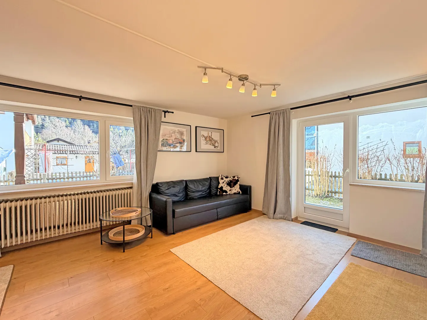 Living room with wood floors, a black sofa, and a light beige rug. Windows with grey curtains look out to a yard.