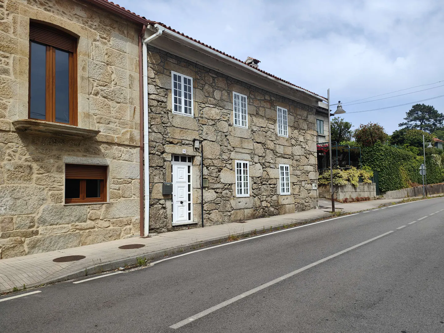 Exterior view of a two-story stone house with white trim windows and a white front door, next to a paved road.
