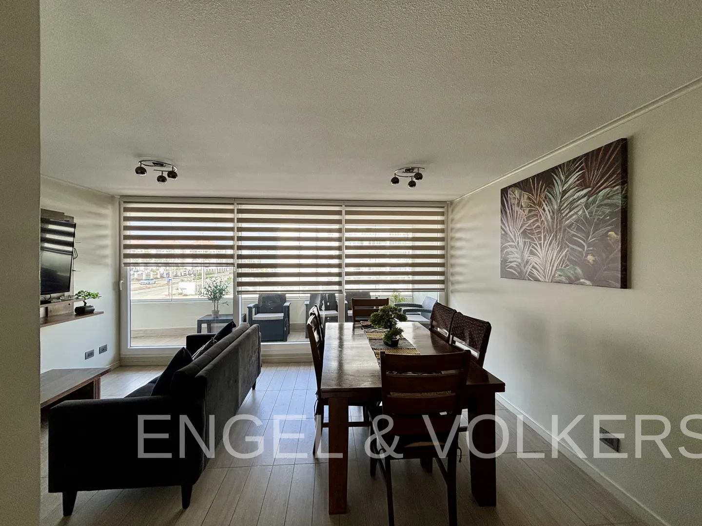 A bright living room with a dark sofa, a wooden dining table, and a large window with blinds. A painting of tropical plants hangs on the wall.