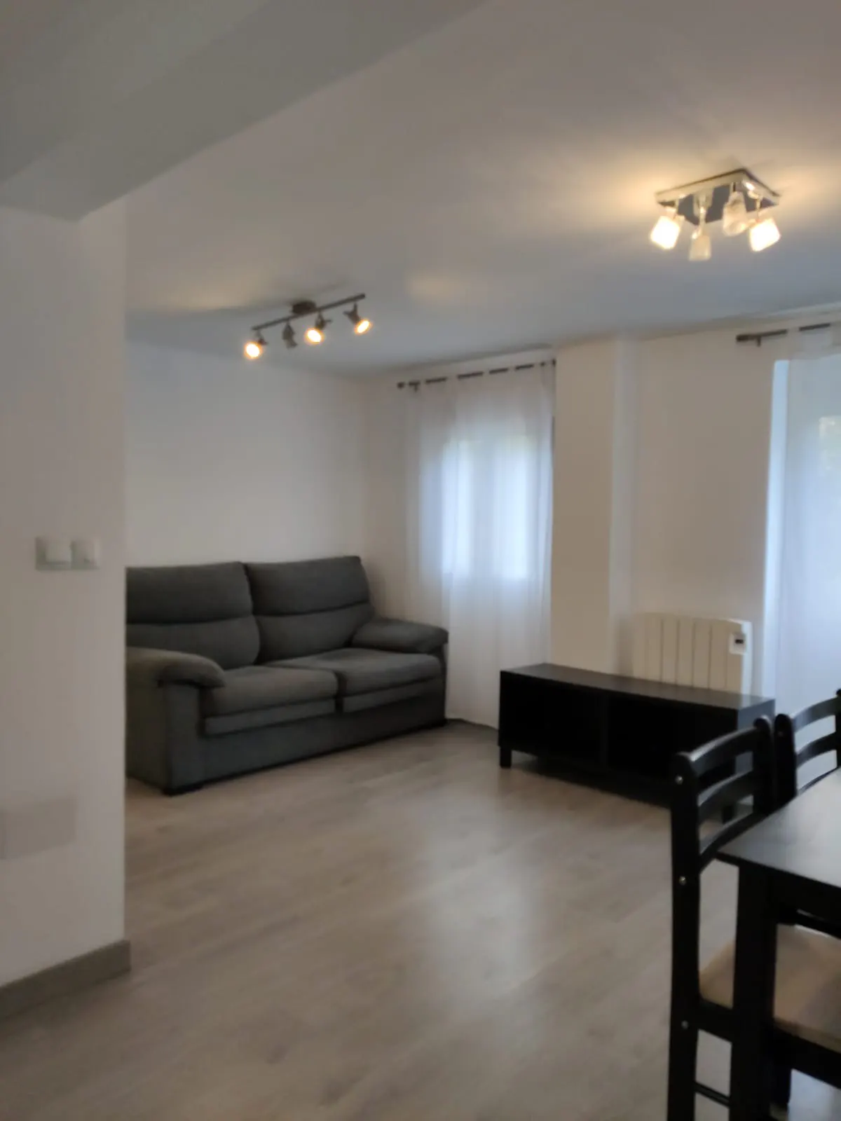 Spacious living room with light wood floors, a grey sofa, a black media console, and a dining area with black chairs.