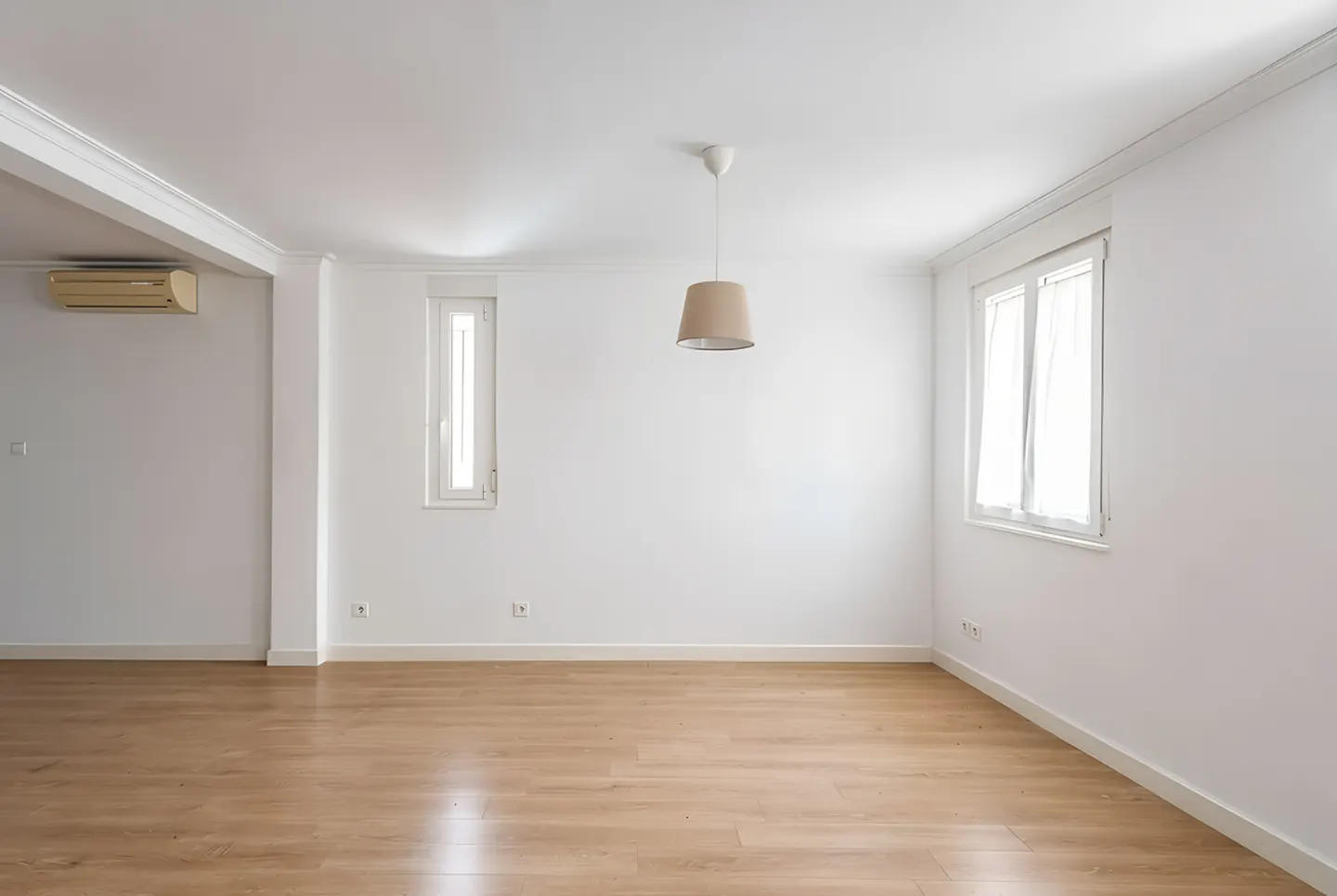 Bright, empty room with light wood floors, white walls, and two windows with white curtains. A beige pendant light hangs from the ceiling.