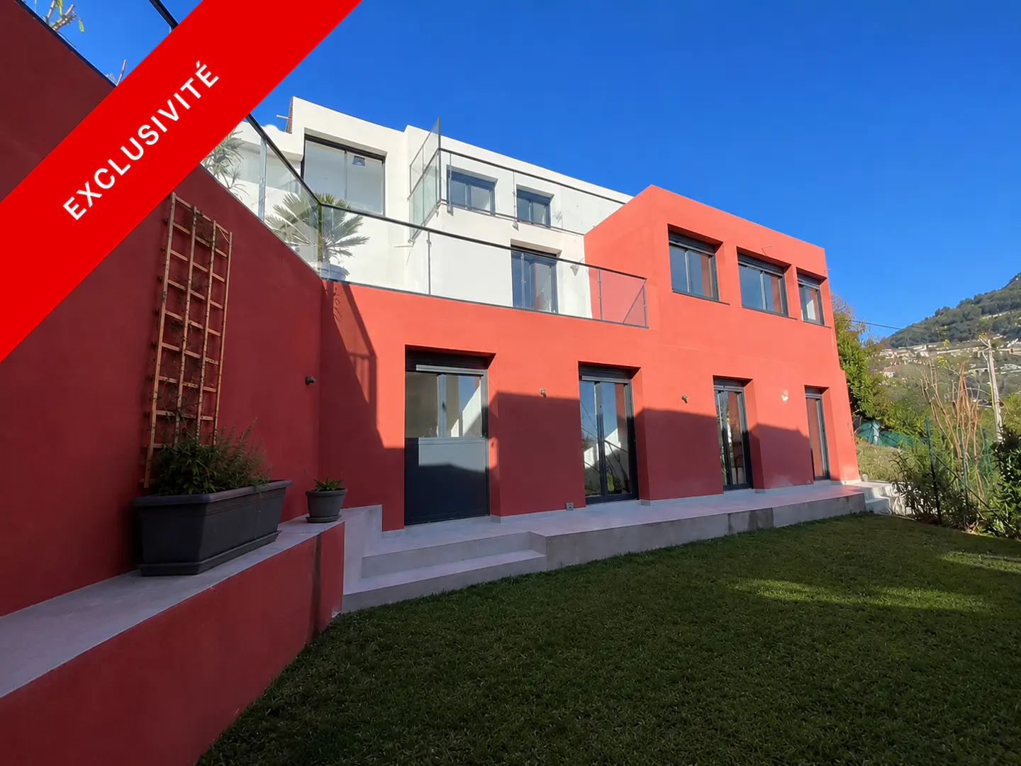 Exterior view of a modern two-story house with red and white walls, a green lawn, and a red "Exclusivité" banner.