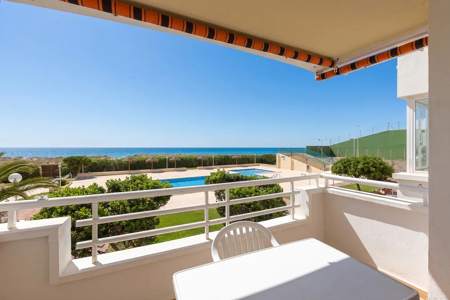 Balcony view of a pool and ocean. White table and chair on the balcony. Blue sky and water. Orange and black awning.
