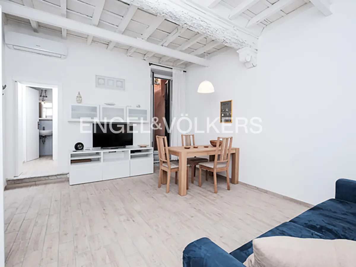 Bright, modern living room with white walls, wood floors, and exposed beam ceiling. A dining table and blue sofa are visible.