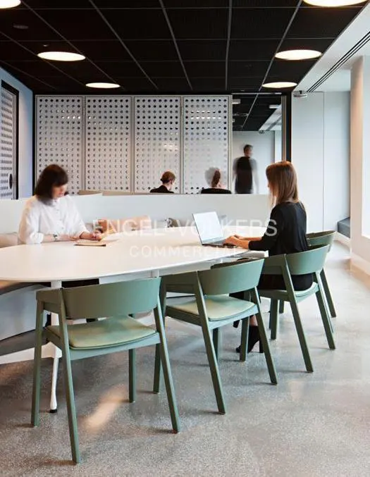Office space with people working at a long white table with green chairs. Black ceiling with round lights.