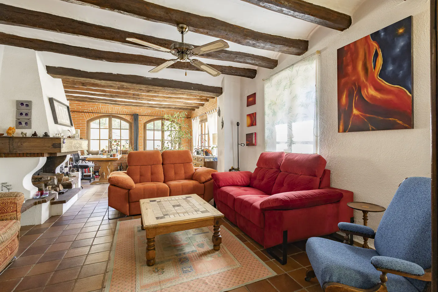 Rustic living room with exposed wood ceiling beams, orange and red sofas, a blue armchair, and a wooden coffee table.
