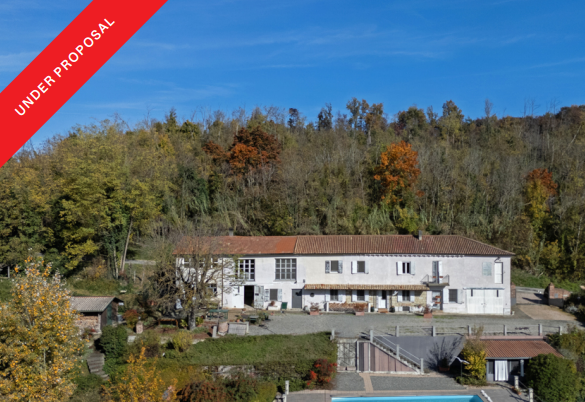 Aerial view of a white farmhouse with a red tile roof, nestled against a wooded hillside under a clear blue sky.