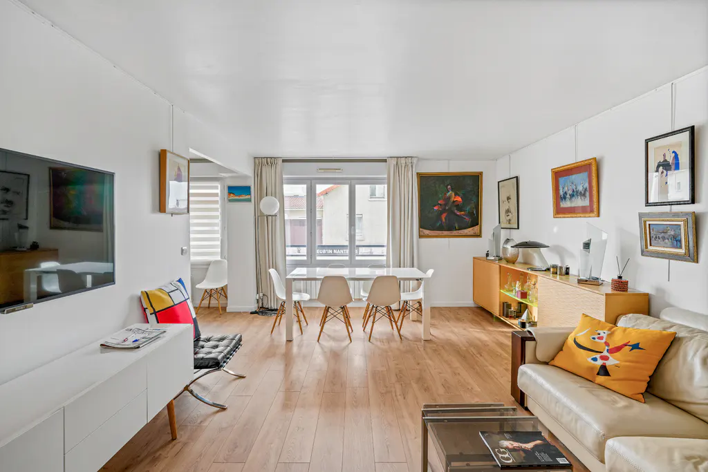 Bright, modern living room with white walls, wood floors, and eclectic art. A white dining table sits near a window. A cream sofa and TV are in the foreground.