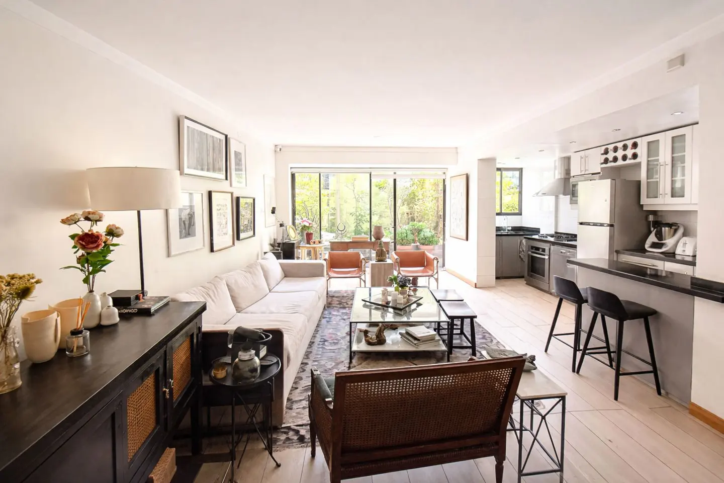 Bright, open-concept living room and kitchen with white walls, light wood floors, and a large sliding glass door to a green outdoor space.