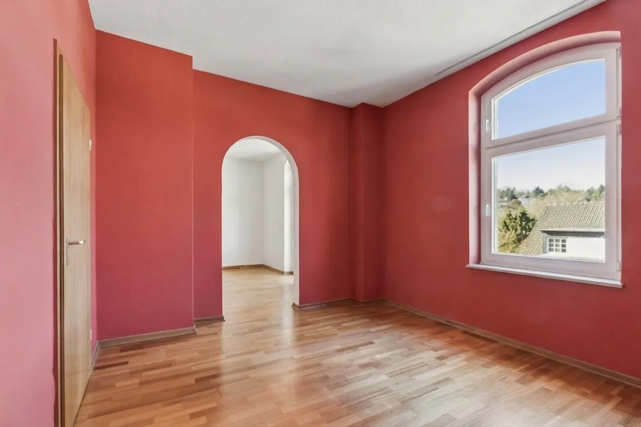 Empty room with red walls, wood floor, arched doorway, and window with view of houses.