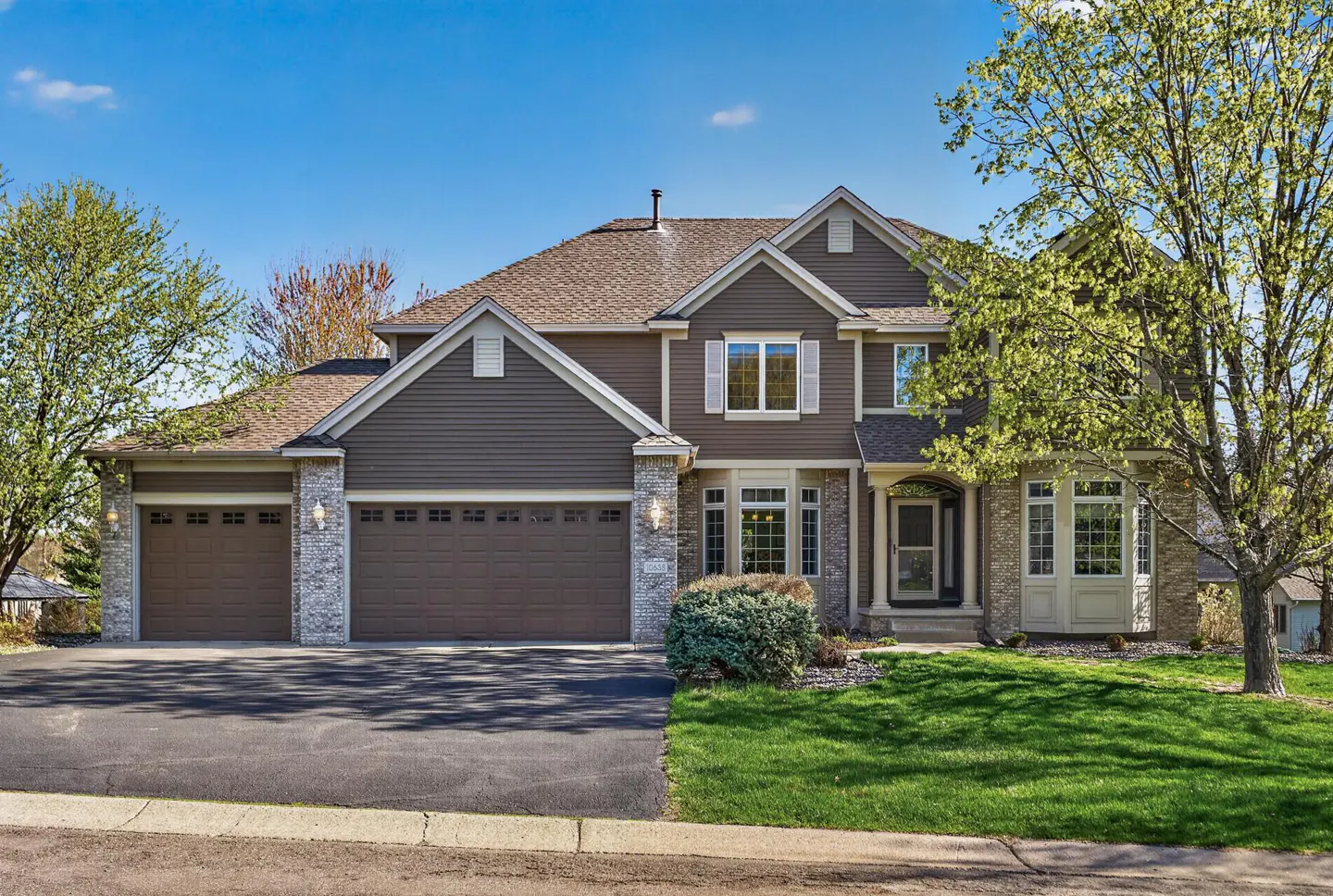 Two-story brown house with a three-car garage, stone accents, and a green lawn under a blue sky.