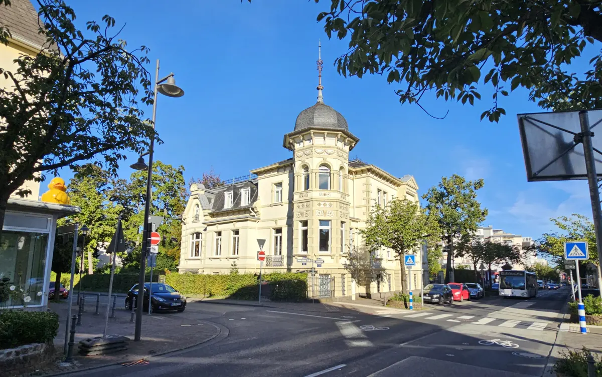 A cream-colored, ornate building with a turret stands at a street corner under a blue sky. Trees and cars line the street.