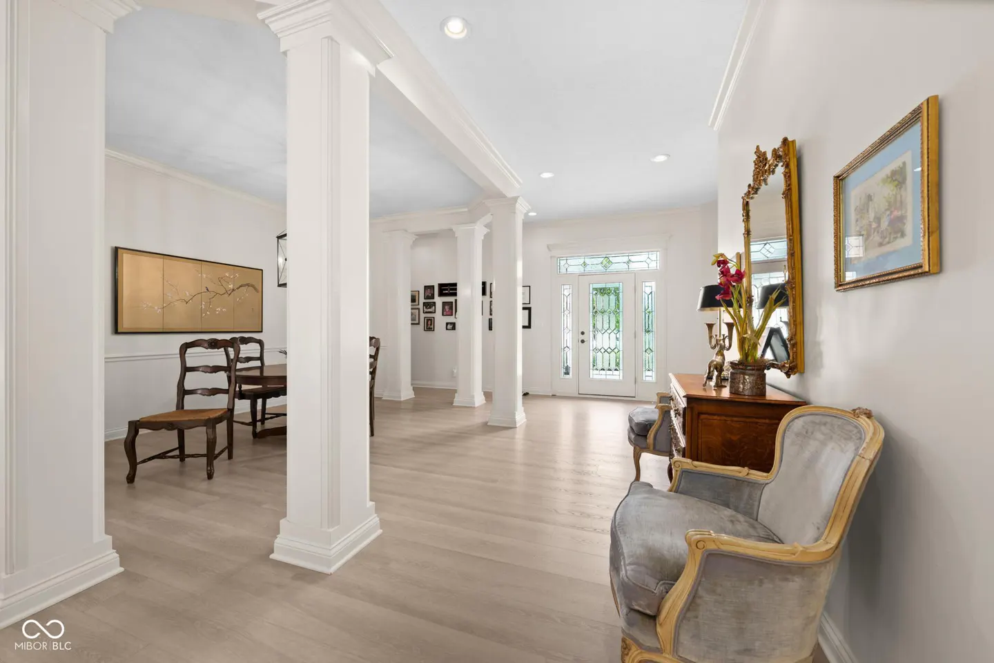 Bright foyer with white columns, light wood floors, and a gray chair. A dining room is visible through the columns.