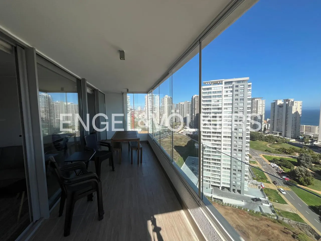 A balcony with glass walls, a wooden table, and chairs overlooks a city with tall buildings and a blue sky.
