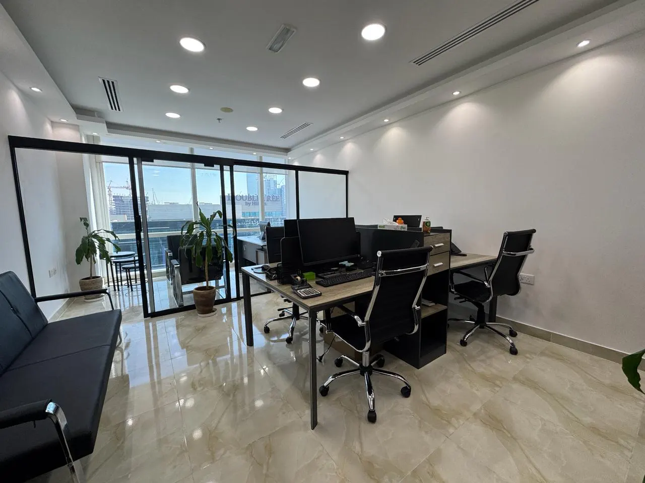 Modern office with a black desk, ergonomic chairs, a black sofa, and large windows overlooking a city skyline.