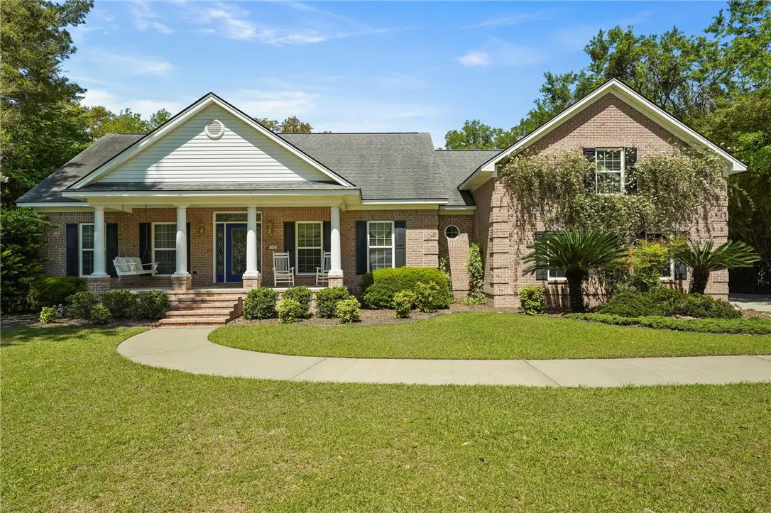 Brick house with white columns and a blue door. A porch swing and rocking chairs sit on the front porch. Green lawn and blue sky.