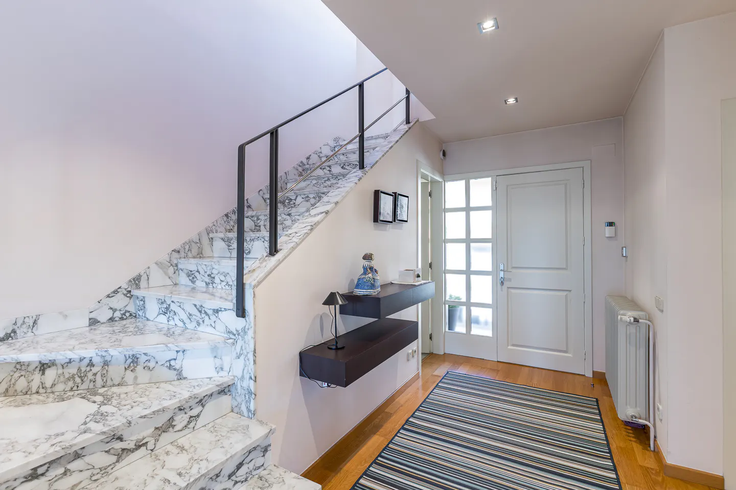 Entryway with marble stairs, black railing, and floating shelves. A striped rug leads to a white front door with glass panels.