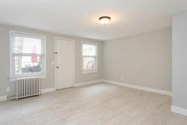 A bright, empty room with light gray walls and tile floors. Two windows with blinds, a white door, and a radiator are visible.