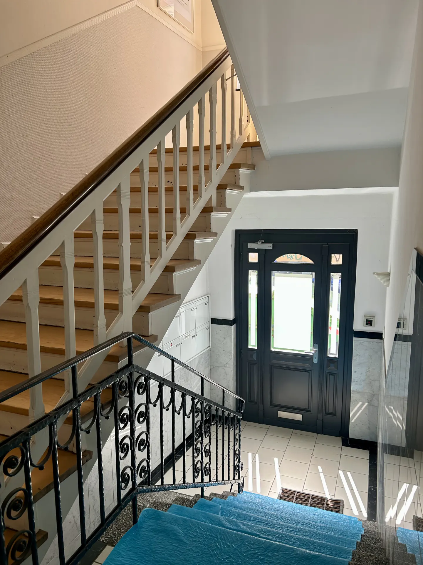 Interior view of a building's staircase with wooden steps, white railings, and a black metal banister. A black door with glass panels is visible in the background.