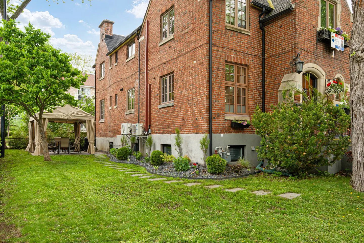 Backyard view of a red brick house with a green lawn, stone path, and a beige gazebo with curtains.