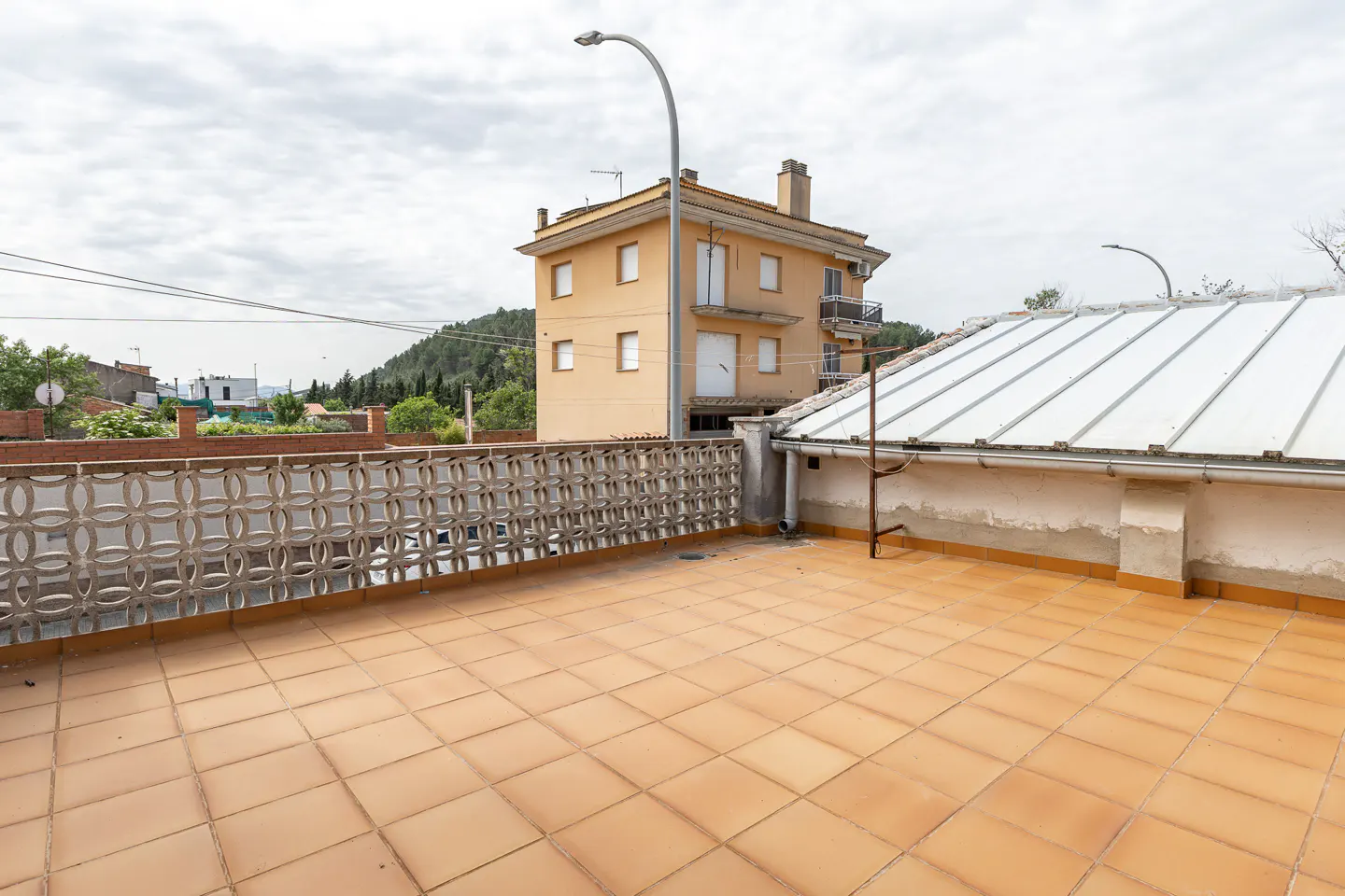Tiled rooftop terrace with patterned concrete railing, overlooking a tan building and cloudy sky.