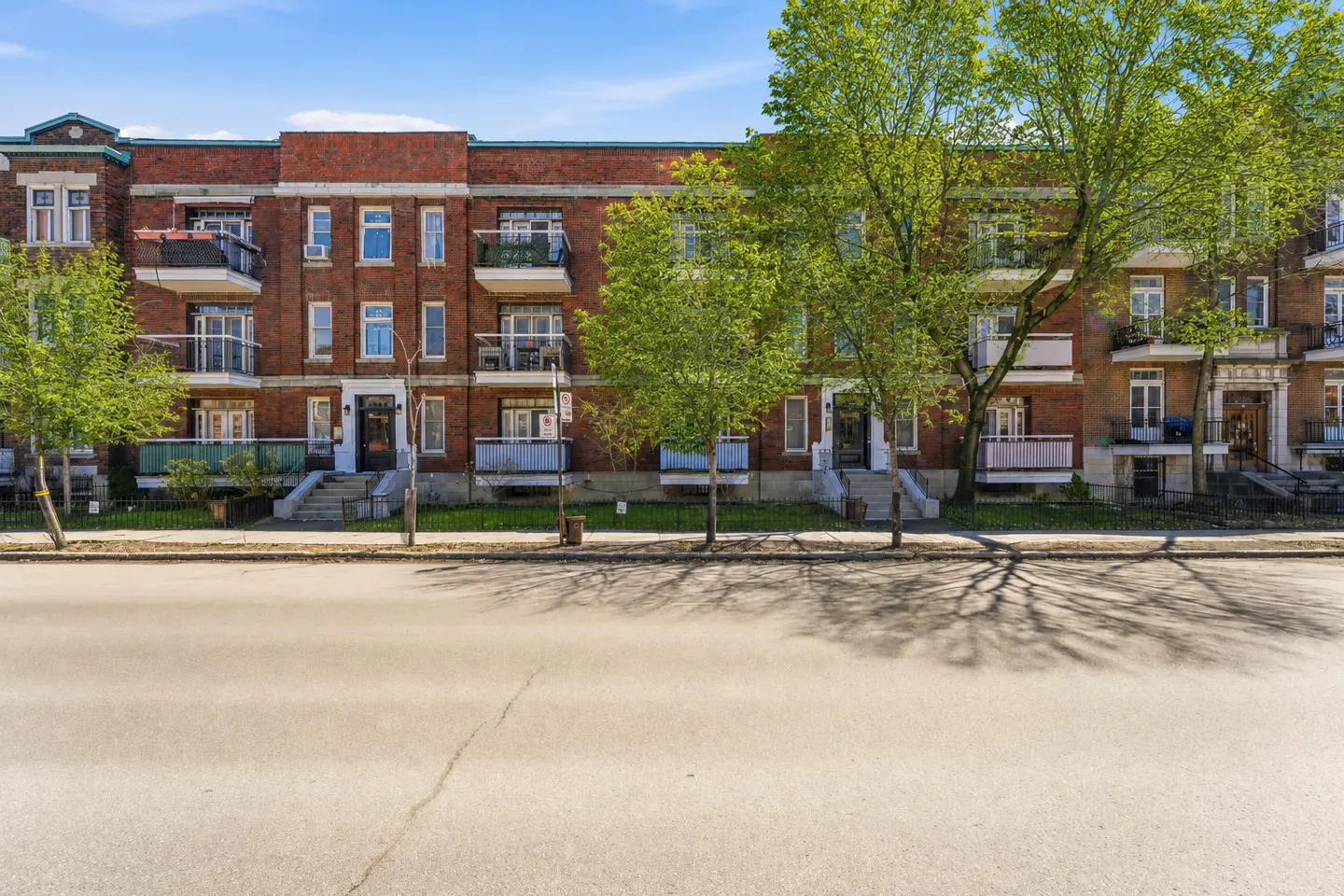Row of red brick apartment buildings with balconies and green trees lining the street under a blue sky.